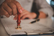 © khunkornStudio - businessman hand stamping approved on certificate document contract in office. Lawyer stamping permit on paperwork at wooden desk.