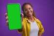 Young smiling African American woman student shows phone with green screen advertising application for self-education and gaining new knowledge through smartphone stands in purple studio. Soft focus