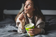 A young woman lies on her bed while eating a pint of pistachio ice cream with spoon. She is lick spoon with ice cream. Eating in bed. Happy beautiful woman resting in her comfortable bed at home. 