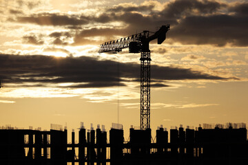 Wall Mural - Silhouette of tower crane and scaffolding of unfinished building at sunrise. Housing construction, apartment block in city on background of dramatic sky with dark clouds