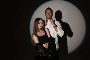 Wall Mural - Beautiful young woman model with a handsome man in fashionable clothes with a white shirt and black leather jacket in the studio on a dark background in a round light