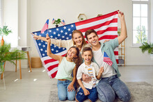 Portrait Of Happy American Family With USA Flag. Mother, Father Their Son And Daughter Sitting With American Flag At Home. Family Celebrating 4th Of July Independence Day