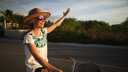 Wall Mural - Closeup side view of happy smiling mature senior woman biking into the sun holding out her arm jubliant, wearing sunglasses hat ethnic attire in a tropical setting with palm trees.