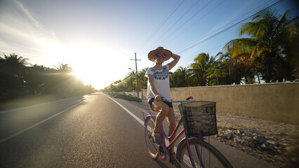 Wall Mural - Low front view of smiling atractive mature senior woman holding her hat, smiling, biking away from the sun, backlit, wearing sunglasses hat, ethnic clothing in a tropical setting.