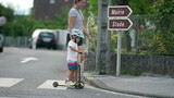 Fototapeta  - mother and child crossing street on sidewalk. Kid wearing helmet riding three wheeled schooter toy transportation on zebra lines with parent