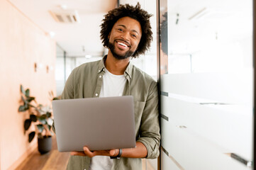 Inspired young african-american businessman looks at camera and smiles cheerfully, standing in modern office and using laptop. Male office employee, freelancer, designer full of new ideas and creative