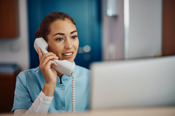 young nurse answering phone call while working on desktop pc at reception desk in hospital.