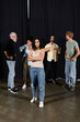multiracial actress standing with crossed arms near interracial theater students and art director talking on background.