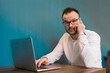 © Andrey - A businessman wearing glasses and a white shirt looks at the camera, talking to his smartphone next to his laptop while sitting at a table in a cafe