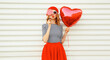 Portrait of happy woman with red heart shaped balloon blowing her lips wearing beret and skirt on white background