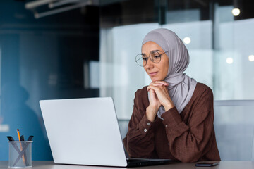Wall Mural - Successful smiling Arab woman in hijab working inside modern office, Muslim woman using laptop at work, business woman satisfied with achievement results typing on computer keyboard.