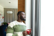 © Wavebreak Media - Thoughtful african american man holding cup of coffee looking out of living room window, copy space