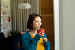 © Wavebreak Media - Thoughtful asian woman standing and drinking coffee, looking out of living room window, copy space