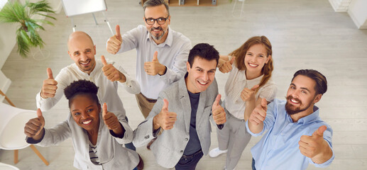 Team of happy smiling business people giving thumbs up together. Joyful diverse male and female workers standing in office, looking up and doing like gestures. Banner, high angle shot. Success concept