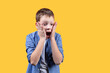 A portrait of a schoolboy showing emotion of shock. He’s holding his cheeks and screaming. He’s looking right and down. Yellow background. Front view. Copy space