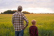 © Acronym - Farmer and his son in front of a sunset agricultural landscape. Man and a boy in a countryside field. Fatherhood, country life, farming and country lifestyle concept.