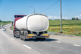 Fototapeta  - Red truck carrying tank semi-trailer on country road. Modern truck pulling white tank trailerswith combustible material through countryside in summer