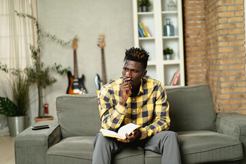 Young African man writing down notes while sitting on sofa at home..