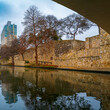 Tranquil San Antonio River Walk footpath, stonewall landscape feature, and boardwalk water reflections with city skyline and barren trees in winter along the canal in Texas, off-season cityscape
