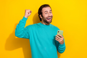 Photo of winning online gambling casino lottery excited guy fist up hold smartphone enjoy get easy internet money isolated on yellow color background