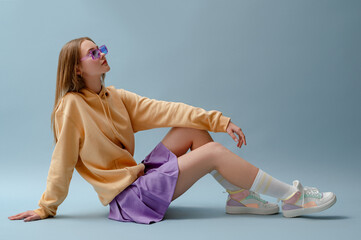 Fashionable confident girl wearing purple sunglasses, pleated mini skirt, yellow hoodie, socks, high top colorful sneakers, sitting, posing on blue background. Full-length studio portrait. Copy space