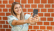 © BooNKer - Young girl taking a selfie - Smiling at the camera - Happy student standing by the wall and using her smartphone