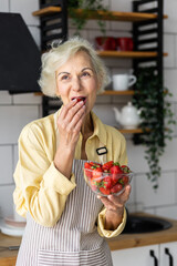 Wall Mural - Close up portrait of attractive senior woman with grey hair cooking healthy food on her kitchen at home. Mature female preparing fresh vegetarian salad with organic ingredients from the market