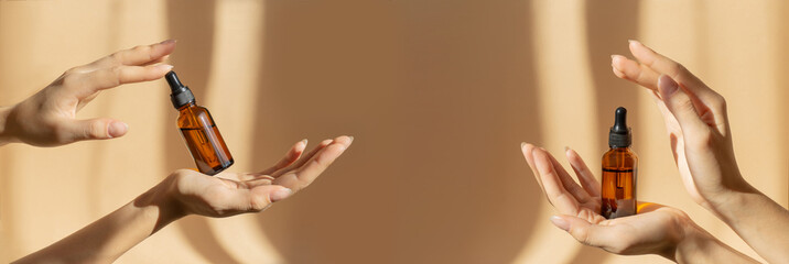 Wall Mural - Two pairs of female hands hold amber glass vials with cosmetic serum on light brown background, in rays of sunlight. Close-up, banner. Branding, mockup concept