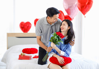 Millennial Asian young romantic lover couple male boyfriend holding red roses bouquet giving surprised to female girlfriend on bed decorated with heart shape balloon on valentine day anniversary