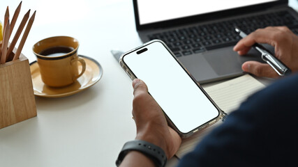 Over shoulder view of man holding smart phone with white empty screen and using laptop on office desk