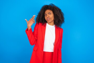 Wall Mural - Unhappy young businesswoman with afro hairstyle wearing red over blue background imitates gun shoot makes suicide gesture keeps two fingers on temples.