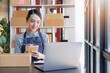 © Natee Meepian - Asian girl startup small business SME, Entrepreneur owner using smartphone or tablet taking receive and checking online purchase shopping order to preparing pack product box. Selling online ideas