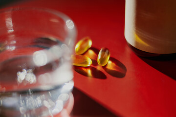 Three yellow dietary supplement pills next to a glass of water, arranged on a red background with copy space.