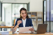 © crizzystudio - Asian businesswoman sitting happily in office holding detailed report paper analyzing data accuracy in financial matters management concept.