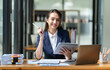 © crizzystudio - Asian businesswoman in a suit sitting holding iPad or tablet in contact See details and discuss business information happily in the office.