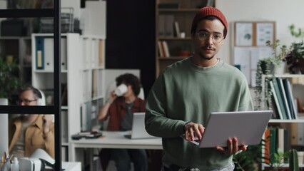 Portrait of young developer looking at camera while working online on laptop with his colleagues in background