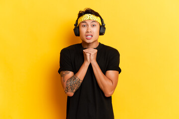 Scared asian man listens music via black wireless headphones finds out something horrible listening audio podcast isolated next to yellow background