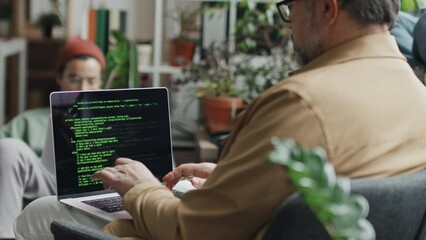 Rear view of mature developer writing codes on computer sitting on armchair with his colleagues in background