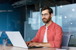 © Liubomir - Serious and focused mature businessman in red shirt wearing glasses looking at camera, young man at work typing on laptop keyboard, programmer developer portrait at work.