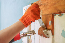 The Process Of Cleaning An Old Wooden Door From Paint Layers. Removal Of The Coating With A Hair Dryer And A Spatula. Worker Hands In Orange Gloves With A Tool. Household Renovation. Copy Space