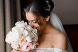 Close view of pretty woman with stylish hairdo and long veil, dressed wedding gown with plunging neckline, enjoying smell of bouquet of flowers during bridal morning