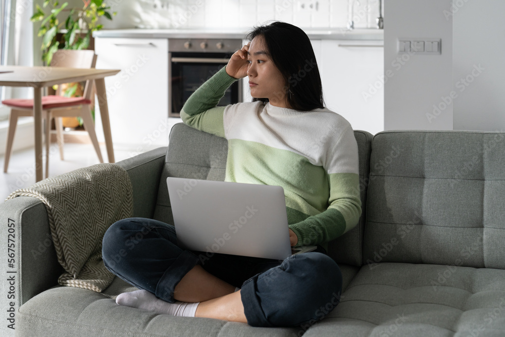 Thoughtful Asian woman freelancer tired sits on sofa with laptop on lap ...