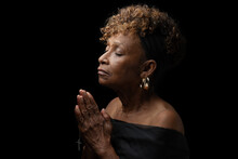 Beautiful Elderly 70  Year Old Christian African American Black Woman Religiously And Fervently Praying To God With Eyes Closed And Hands Together Holding A Cross