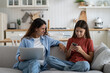 © DimaBerlin - Young smiling mother talking to teen daughter child about being online, modern family using digital devices while resting on sofa at home. Mom chatting speaking with kid about internet safety