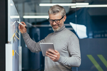 Mature man inside office near whiteboard with colored notes writing down strategy and business plan, gray haired businessman with tablet thinking at work inside office smiling happy with result.