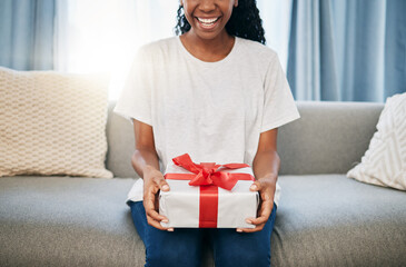 Wall Mural - Present, box and woman on a sofa in the living room with a giving gesture for celebration or event. Bow, wrapping paper and African female with a present for christmas, birthday or holidays in house.