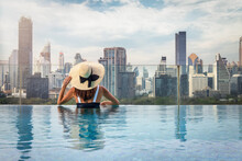 A Woman With Hat Enjoys The Elevated View Over The Urban Skyline Of Bangkok, Thailand, From A Swimming Pool