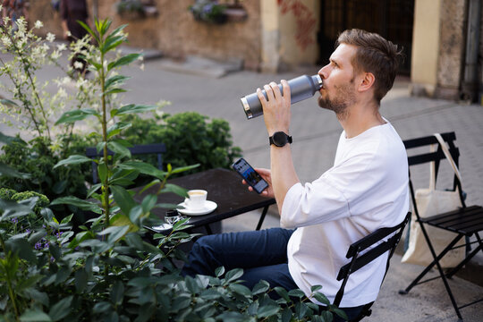 In a sustainable city, a man in a white shirt drinks from a reusable metal bottle at a green street cafe while using his smartphone. A coffee cup sits on the table as he enjoys a 
