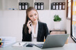 © NINENII - Asian woman  talking mobile phone and working on laptop and papers on table in her office.