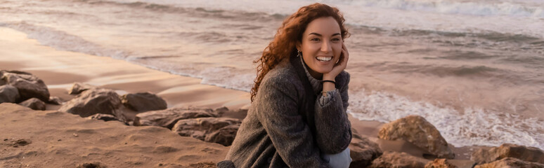 Wall Mural - Cheerful young woman looking away while sitting on beach during sunset, banner.